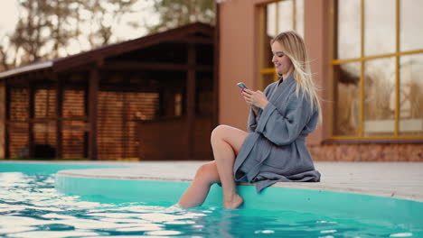 charming-young-blonde-woman-is-relaxing-near-warm-outdoor-pool-in-cold-weather-chatting-online-by-smartphone